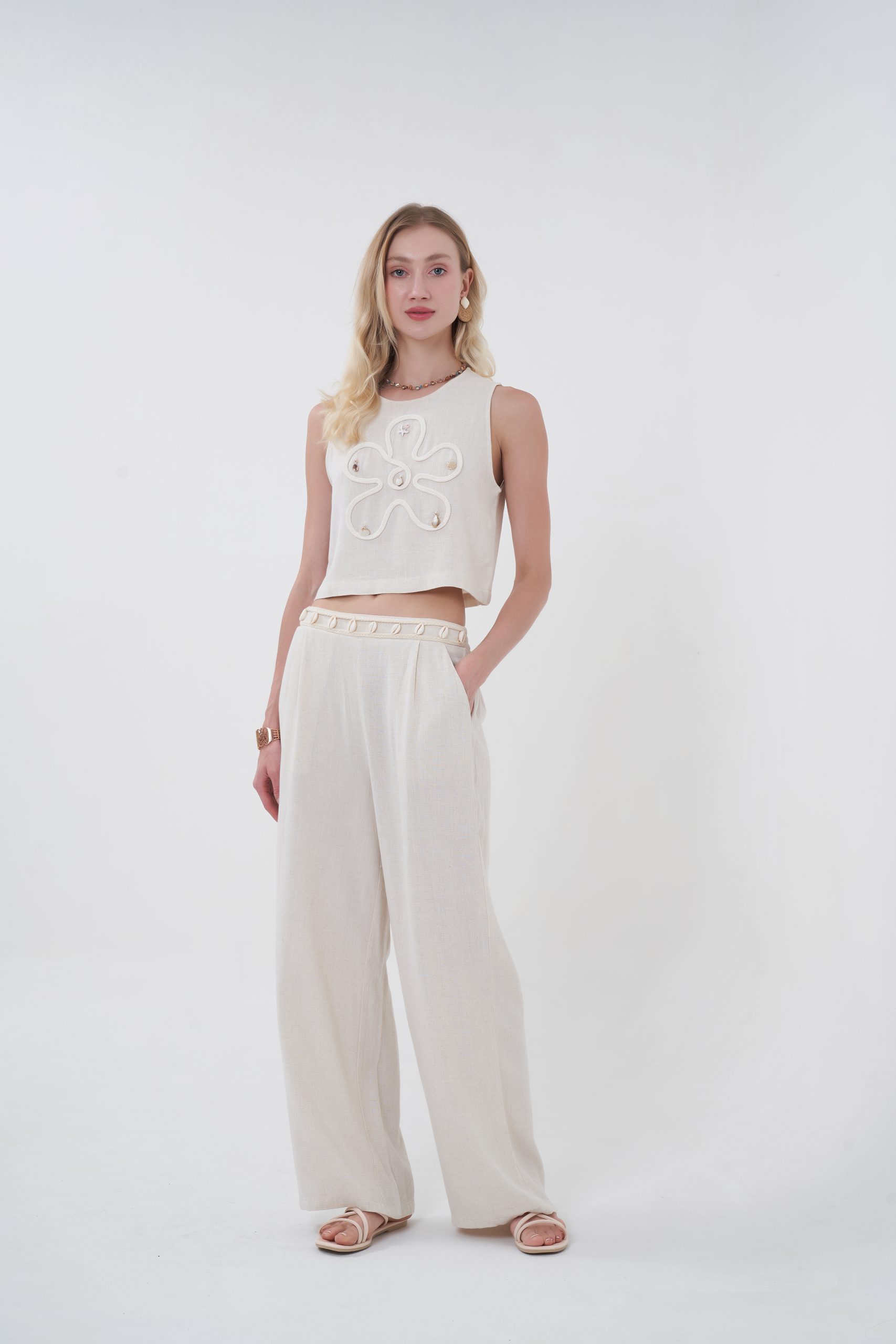 A warm-weather statement piece, this shell embellished crop top features a textured woven fabric in a soft beige tone, designed with a relaxed sleeveless silhouette and cropped length for effortless styling. The front is delicately adorned with shell accents arranged in an organic motif, adding a subtle coastal touch while keeping the look modern and elevated. Lightweight and breathable, it’s perfect for sunny days, vacations, or relaxed resort evenings.